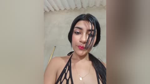 Video of a young woman with long black hair, wearing a black bikini top, standing in a shower with beige tiles.