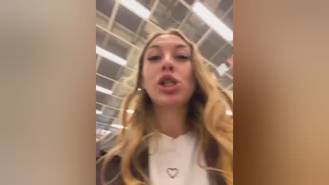A video of a young woman with long, wavy blonde hair and light skin, wearing a white shirt with a heart pendant, posing indoors with a ceiling grid in the background.