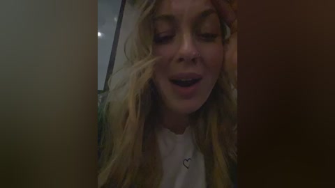 Video of a blonde woman with closed eyes, leaning against a wooden surface, wearing a white shirt.