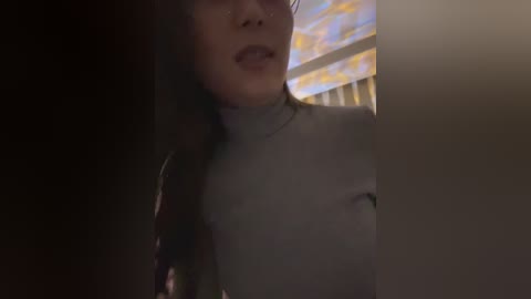 Video of a woman with light skin and long brown hair, wearing a light gray turtleneck sweater, partially obscured by shadow. Background features a window with yellow curtains.