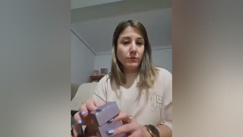 Video of a young woman with light skin and straight, shoulder-length blonde hair, wearing a white t-shirt, holding a small, lavender-colored box in a living room with white walls and beige furniture.