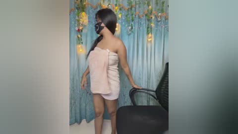 Video of a woman with medium brown skin, black hair, and a black mask, wrapped in a towel, standing in a room with blue curtains and fairy lights.