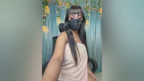 Video of a young woman with long black hair, wearing a pink towel and a black face mask, seated in a teal room adorned with hanging flower lights.