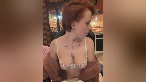 A video of a fair-skinned woman with short red hair, wearing floral lingerie, tattoos on her chest, sitting in a dimly lit room with a fish tank and a kitchen in the background.