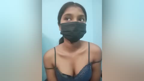Video of a young woman with medium-dark skin, wearing a black mask and a dark blue lace bra, standing against a light blue wall.