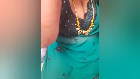 Video of a woman wearing a teal saree with black lace trim, adorned with gold jewelry. Background blurred, focusing on her attire and necklace.