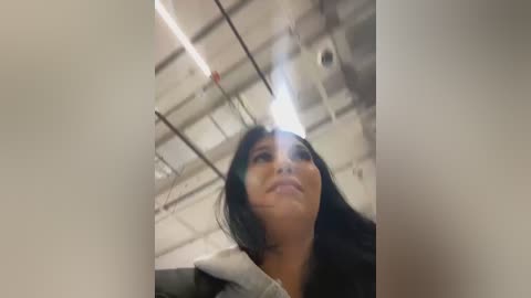 Video of a woman with long black hair and a serious expression, taken from a low angle, showing her from the neck up. The background features a white ceiling with exposed ductwork.