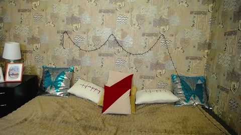 Video of a cozy, vintage-style bedroom with beige wallpaper, featuring a bed with a beige bedspread, two teal pillows, a red and white geometric pillow, and a framed photo on a black nightstand.