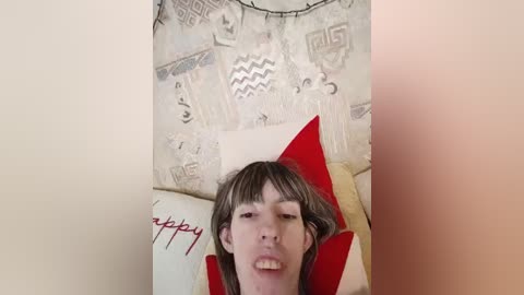 A video shows a young man with straight, light brown hair lying on a bed with white and red pillows, against a beige, patterned wall. He has a neutral expression.