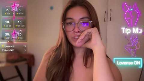 Video of a topless woman with light skin and long brown hair, wearing glasses, lying on a bed. Background includes a TV showing \"Tip Max\" and \"Love Me\" game menus.