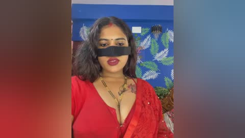 Video of a South Asian woman with dark skin, long black hair, and a red sari, wearing a black blindfold and red lipstick, in a room with blue walls and a leaf-patterned tapestry.