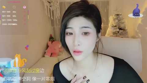 A video of a young East Asian woman with fair skin and dark hair, wearing a black top, in a cozy room with plush toys, a bed, and a plush teddy bear.