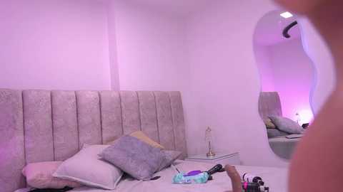 Video of a minimalist, softly lit bedroom with a tufted, gray upholstered headboard, a round mirror, and a sleek white nightstand holding a lamp and jewelry.