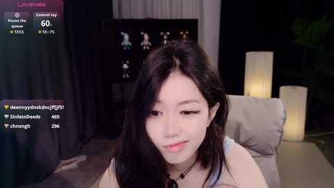 Video of a young Asian woman with long black hair, light skin, and makeup, sitting on a sofa in a dimly lit room. She has a neutral expression.
