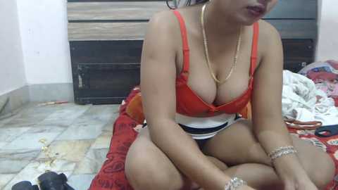 Video of a dark-skinned woman with medium-sized breasts, wearing a red lace bra and black shorts, sitting on a red blanket in a cluttered room with wooden furniture and a tiled floor.