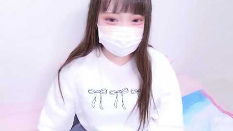 Video of an East Asian woman with long brown hair, wearing a white face mask and white hoodie with a ribbon graphic, sitting against a pastel-colored background.