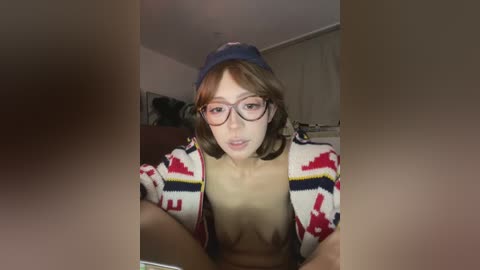 Video of a fair-skinned woman with shoulder-length brown hair, wearing glasses, a blue cap, and an open, patterned cardigan. She's topless, with visible breasts, in a dimly lit room with a bed and a TV in the background.