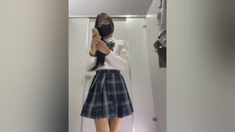 Video of a slim, light-skinned Asian woman with long black hair, wearing a white blouse, plaid skirt, and black mask, standing in a dimly lit, narrow hallway with a closed door and hanging clothes.