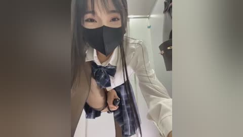 Video of an Asian woman in a revealing schoolgirl outfit, featuring a plaid skirt, white jacket, and black mask, with exposed breasts, standing in a dimly lit room.