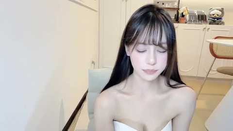 Video of a young Asian woman with long black hair, wearing a strapless white dress, sitting in a modern kitchen with white cabinets and a table.
