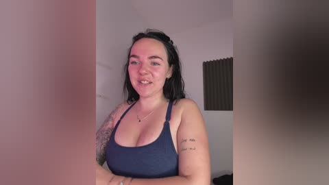 A video of a smiling, pale-skinned woman with dark hair, wearing a low-cut, dark blue tank top, tattoos on her arms, in a dimly lit room with a window with blinds.