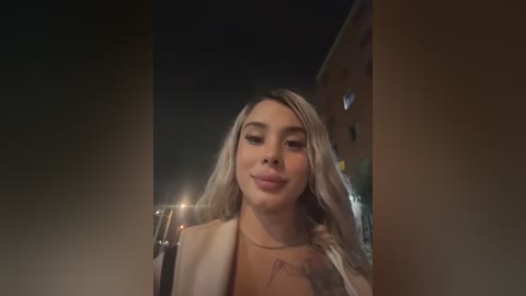 Video of a fair-skinned woman with long, wavy blonde hair, wearing a beige jacket and a necklace, smiling slightly, taken at night with a dimly lit urban background.
