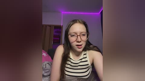Video of a young Caucasian woman with long brown hair, wearing glasses, a black and white striped tank top, and a neutral expression. Background includes a pink pillow, a wooden wardrobe, and purple LED lights.