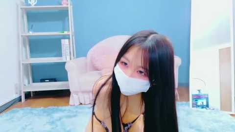 Video of an East Asian woman with long black hair, wearing a white surgical mask, kneeling on a light blue rug in a pastel-colored living room with a white bookshelf and pink chair.