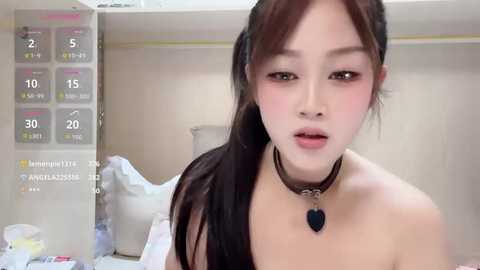 Video of an East Asian woman with light skin, dark hair, and wearing a black choker necklace, standing in a hospital room with a temperature monitor and a bed visible in the background.