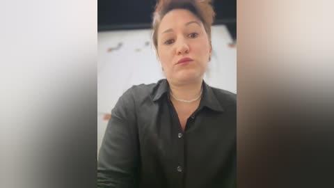 Video of a middle-aged woman with light skin and brown hair, wearing a black button-up shirt, and a pearl necklace, looking serious, with a blurred background.