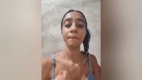 Video of a young woman with medium brown skin and dark hair, wearing a light blue bra, standing against a textured gray wall. She has a neutral expression and is slightly off-center.