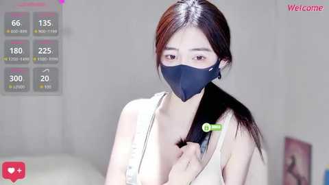 A video of an East Asian woman with fair skin, long dark hair, and a black face mask, wearing a white tank top. She's indoors with a simple background.