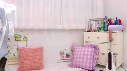 A video of a whimsical, pastel-themed bedroom corner featuring a white bed with pink and purple checkered pillows, a white dresser with colorful toys, and a potted plant.