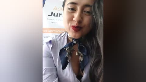 Video of a woman with long, dark hair, wearing a purple blouse with a deep V-neck and a blue scarf with silver accents, smiling against a blurred background.
