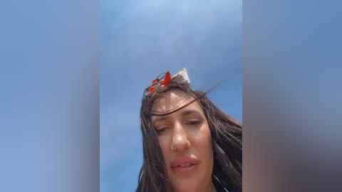 A video of a young woman with long, wet hair, wearing a red headband, looking down under a clear blue sky.