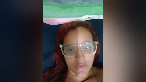 Video of a woman with long, reddish-brown hair, wearing clear glasses, lying on a dark bed. A green and white pillow is visible above her head.