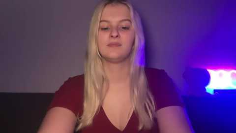 Video of a young Caucasian woman with long, straight blonde hair, wearing a deep burgundy V-neck top, sitting indoors with a dim, purple-lit background.