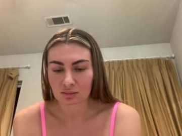 Video of a young woman with light skin and straight, shoulder-length brown hair, wearing a pink tank top. She appears to be applying makeup. Background features beige curtains and a white ceiling.