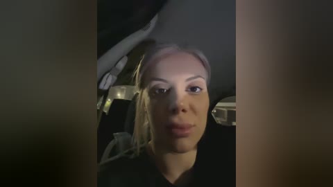 Video of a woman with light skin and blonde hair, wearing a black hoodie, sitting in a car. The dimly lit interior has a black steering wheel and blurry background.