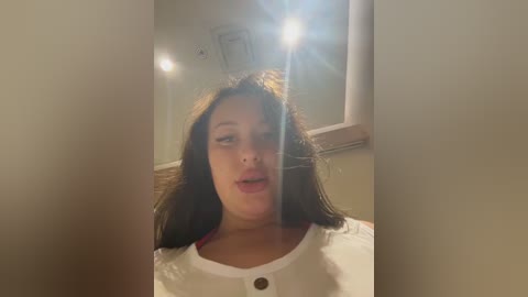 Video of a woman with long brown hair and light skin, wearing a white top, standing in a dimly lit room with soft lighting. She has a neutral expression.