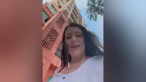 Media: Video of a young woman with long, dark hair, wearing a white top, standing in front of a pink building with balconies and palm trees in the background.
