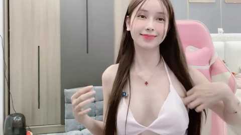 Video of a young, fair-skinned Asian woman with long brown hair, wearing a pink halter top, sitting in a pink gaming chair. Background features light wood cabinets and a blue cushioned bench.