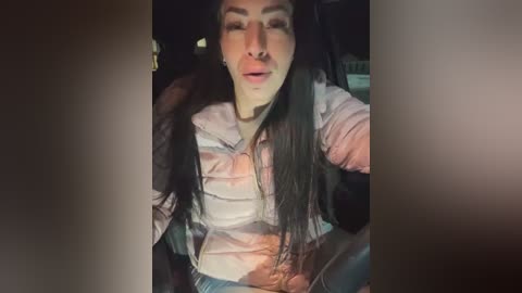 Video of a woman with long dark hair, wearing a light pink puffer jacket, sitting in a car at night. Her face is partially obscured by the reflection of the car window.