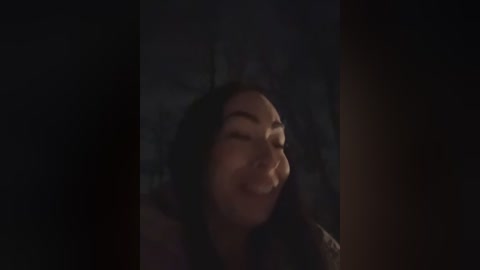 Video of a smiling woman with long dark hair, wearing a dark top, partially obscured by a dimly lit background.