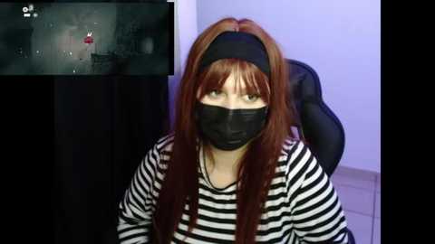 Video of a woman with long auburn hair, wearing a black face mask, black headband, and black-and-white striped shirt, sitting in a black chair in a dimly lit room with a TV screen showing a video feed.