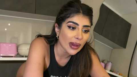 Video of a woman with long dark hair, wearing black lipstick, and a black top, leaning over a counter with pink and white kitchen items in the background.