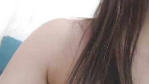 A close-up video of a woman's bare shoulder and long brown hair, with a blurred background of a white wall and a teal pillow.