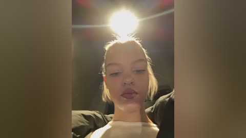 Video of a young woman with light skin and blonde hair, wearing a black jacket, looking contemplative in a dimly lit room with a bright light source above.