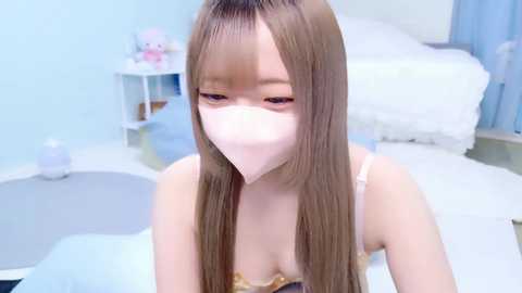 A video of an Asian woman with long, straight brown hair, wearing a pink face mask and a light pink bralette, sitting in a pastel-colored bedroom with a white bed, soft lighting, and minimalistic decor.