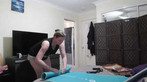 Video of a woman in a black sports bra and shorts rolling out a yoga mat in a cozy, dimly lit living room. The background features a TV, a wicker screen, and hanging clothes.